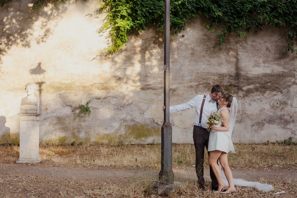 Groom leaning on a lamppost while bride embraces him beneath leafy wall.