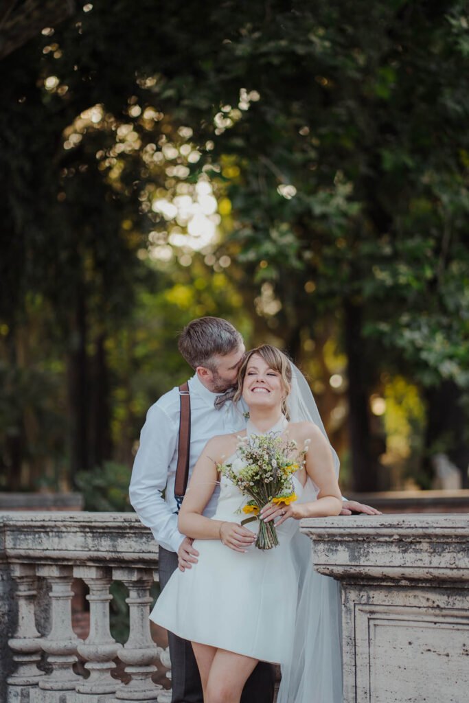 Bride smiling while groom hugs her near a stone balustrade in Rome.