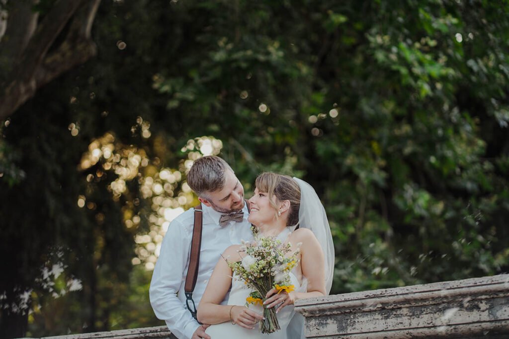 Bride and groom looking at each other while sitting on a stone wall.
