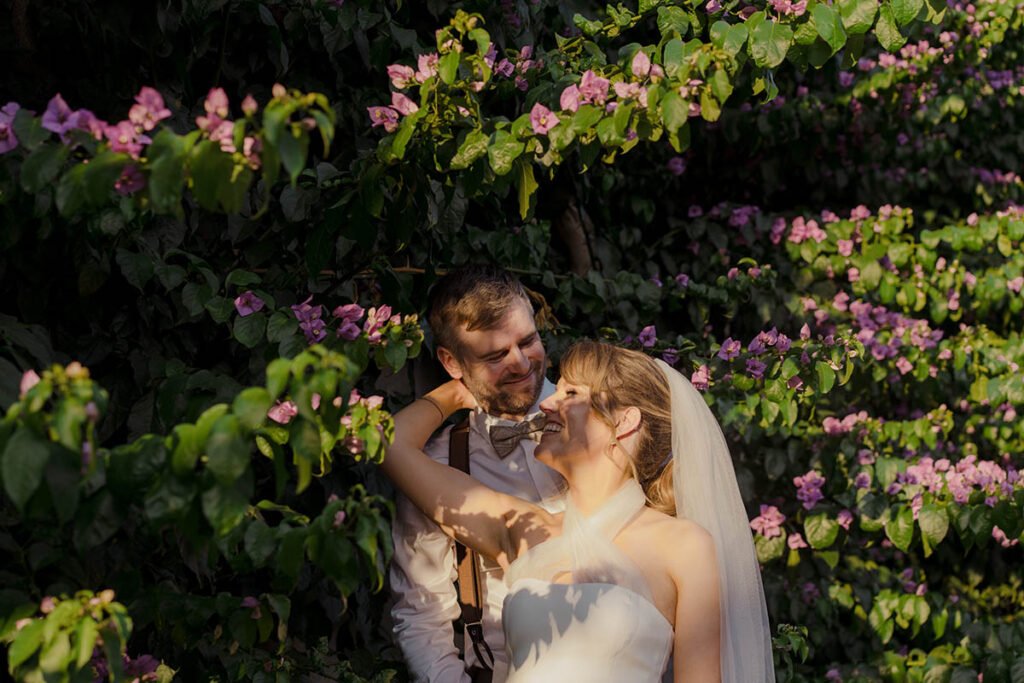 Groom hugging the bride from behind under purple flowers.