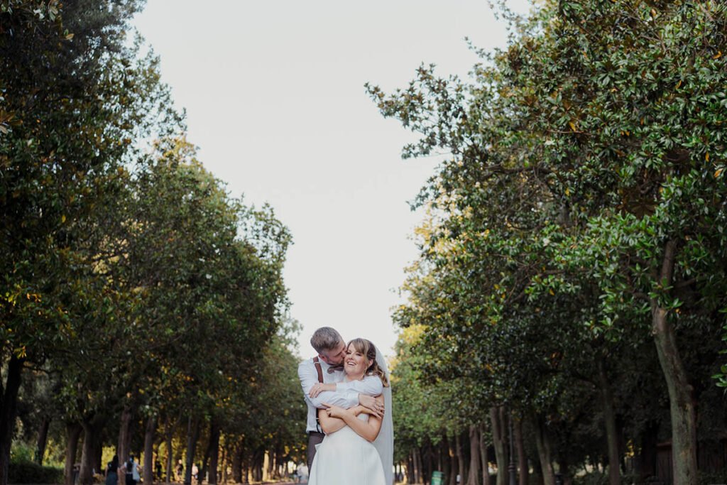Groom holding the bride from behind on a tree-lined avenue in Villa Borghese.
