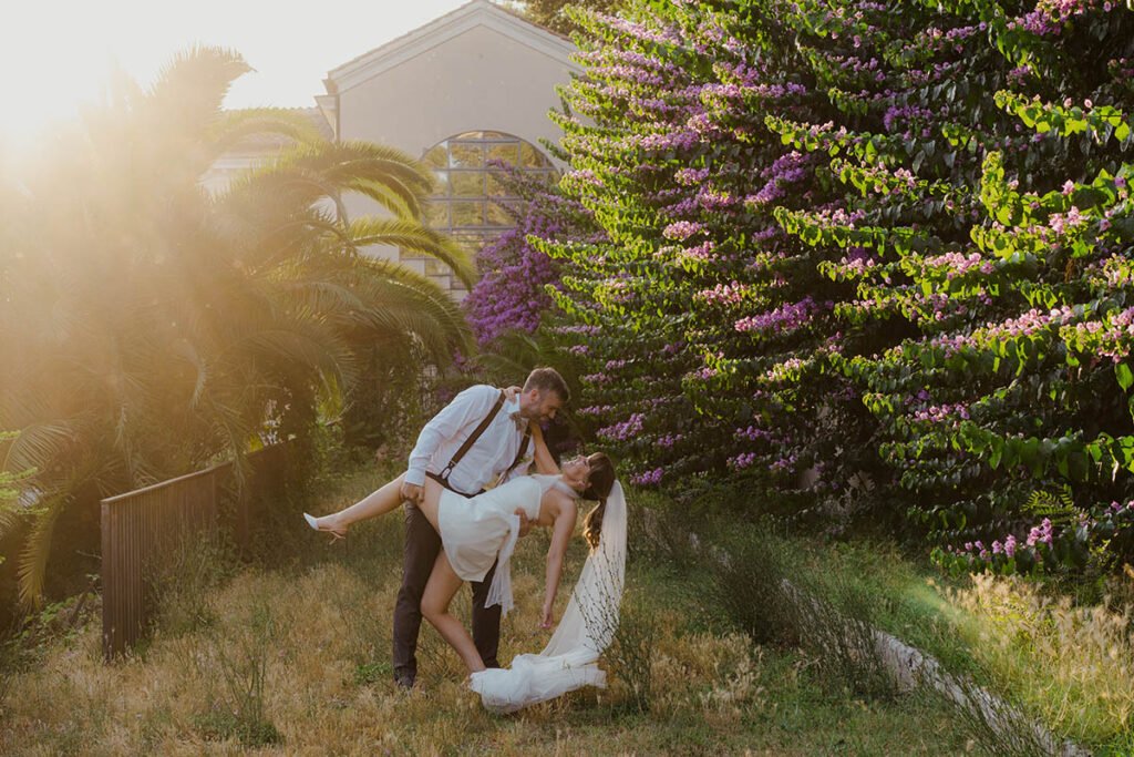 Groom dipping the bride while surrounded by vibrant purple flowers and palms.