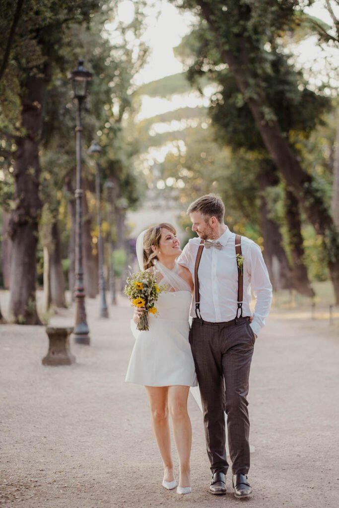 Bride and groom walking hand in hand along a shaded path in Villa Borghese.