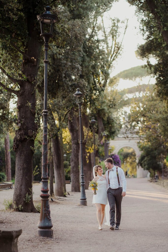 Couple walking hand in hand under tall trees with Rome skyline behind.