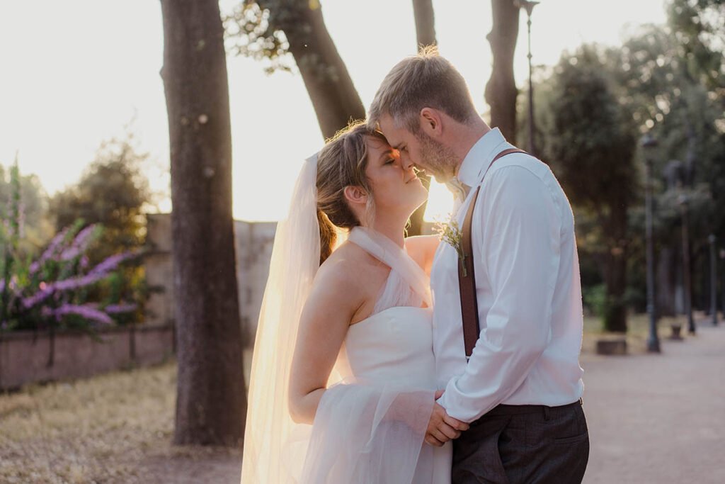 Bride and groom kissing at sunset among tall trees in Villa Borghese.