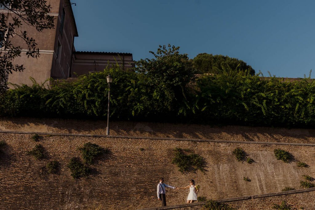 Bride and groom walking below a Roman wall with vines above.