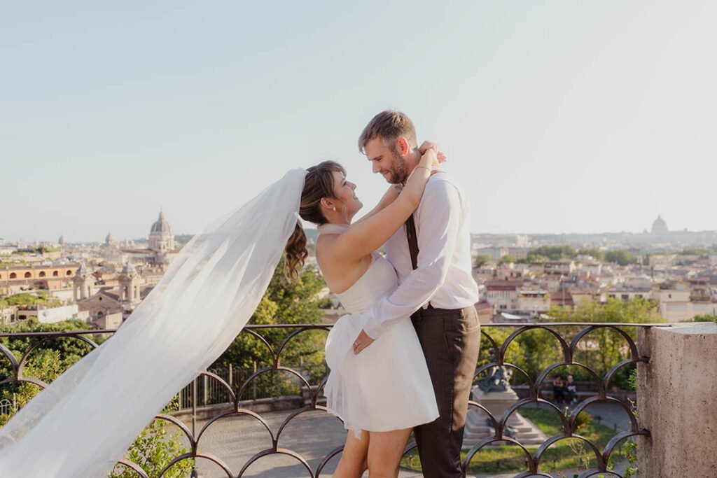 Bride smiling with veil in the wind as the groom lifts her against a city view.