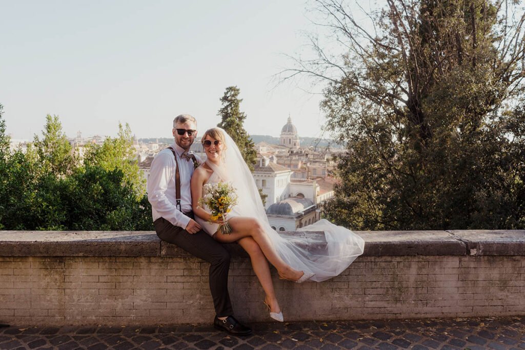 Bride sitting on a wall with groom behind her and bouquet in hand.
