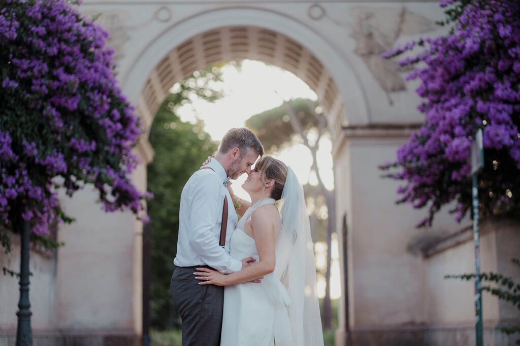 Couple standing close under a stone archway covered with purple flowers.