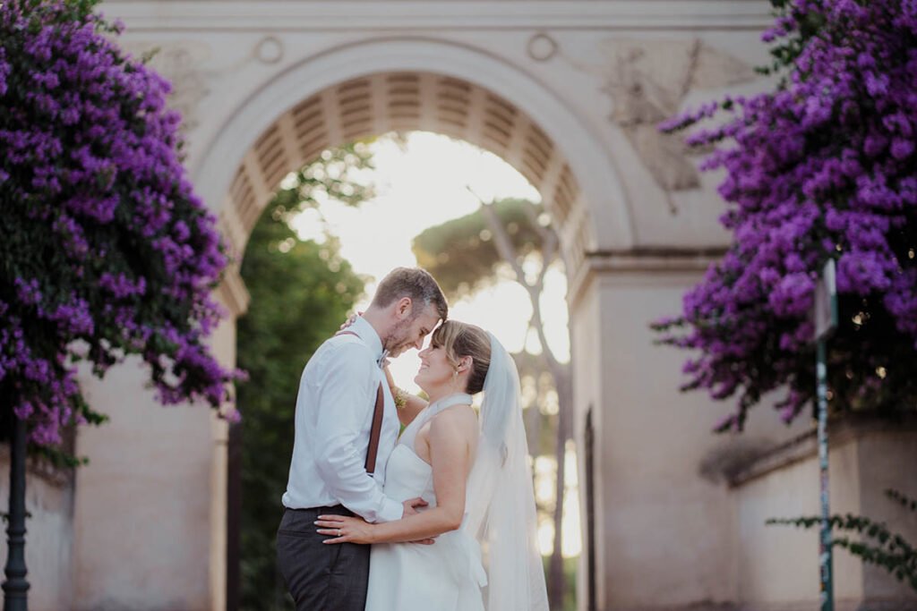Couple sharing a kiss under a Roman archway with purple flowers.