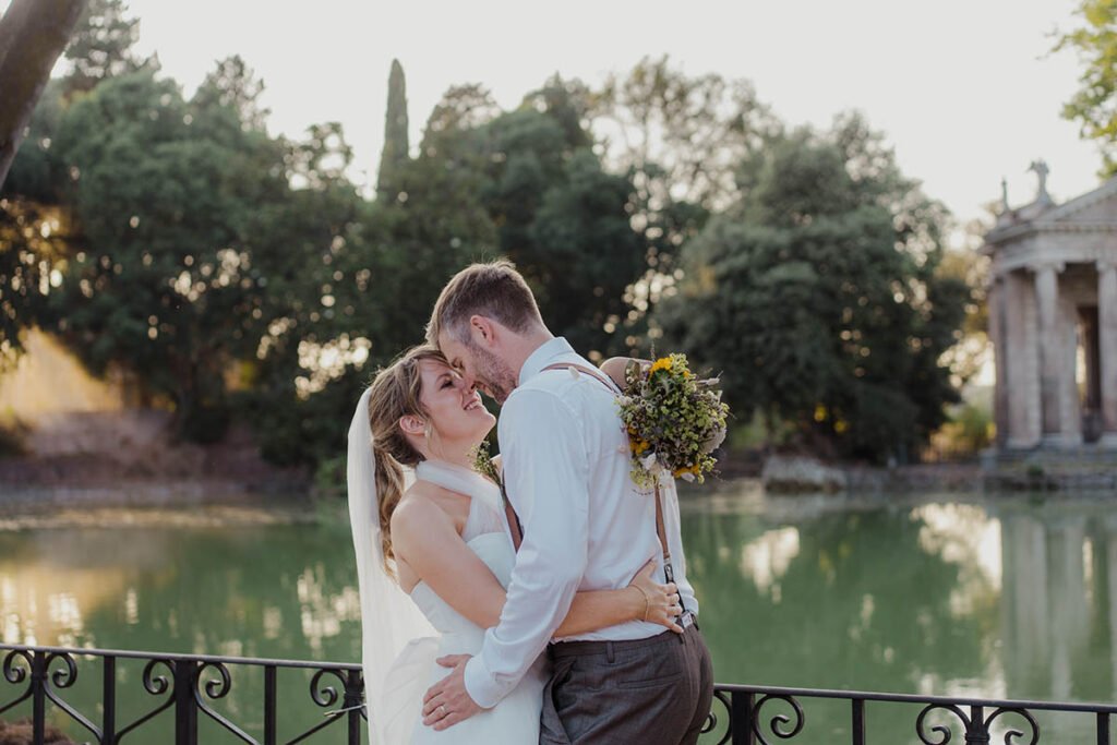 Couple embracing near a calm lake in Rome's Villa Borghese park.