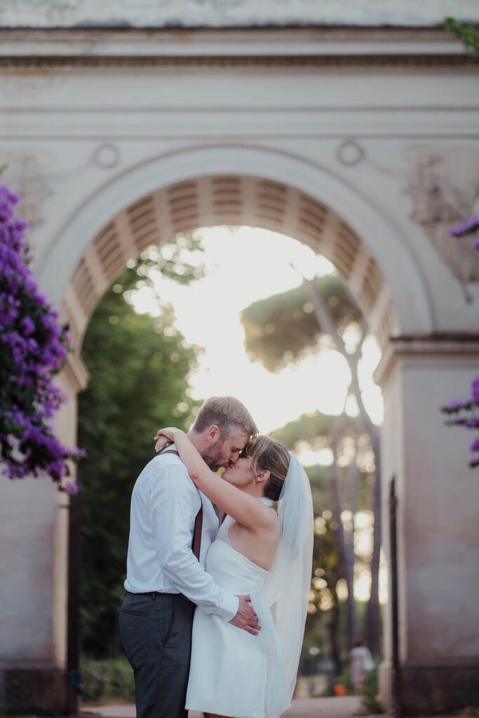 Couple kissing under a stone archway with sun flaring behind them.